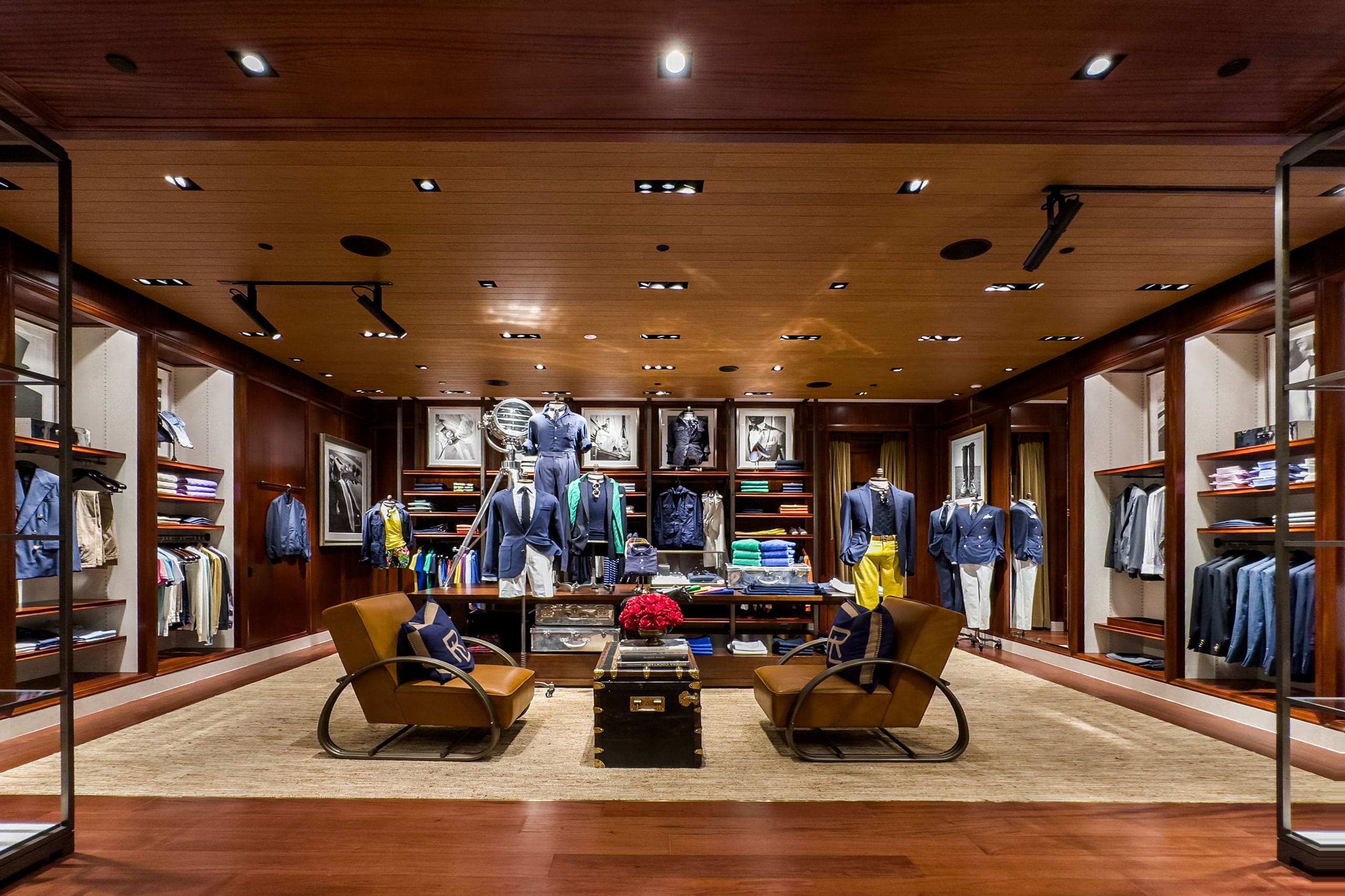 Upscale clothing store interior with wooden walls and shelves displaying neatly arranged shirts, jackets, and accessories. Two leather chairs face a table with red flowers, and mannequins are dressed in stylish outfits.