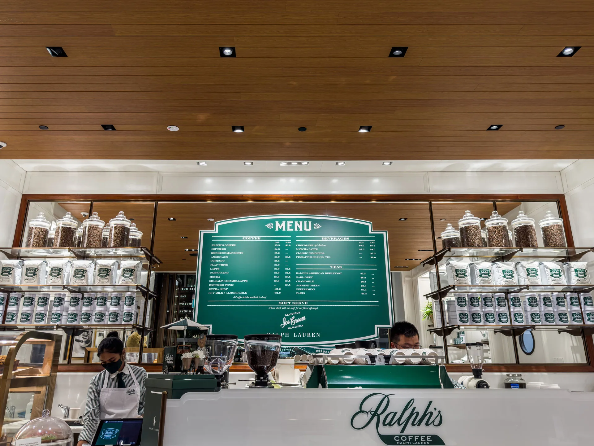 A coffee shop counter with two staff members, one wearing a mask, in front of a green menu board and shelves lined with jars and tins labeled Ralphs Coffee. The shop has a wooden ceiling and elegant decor.
