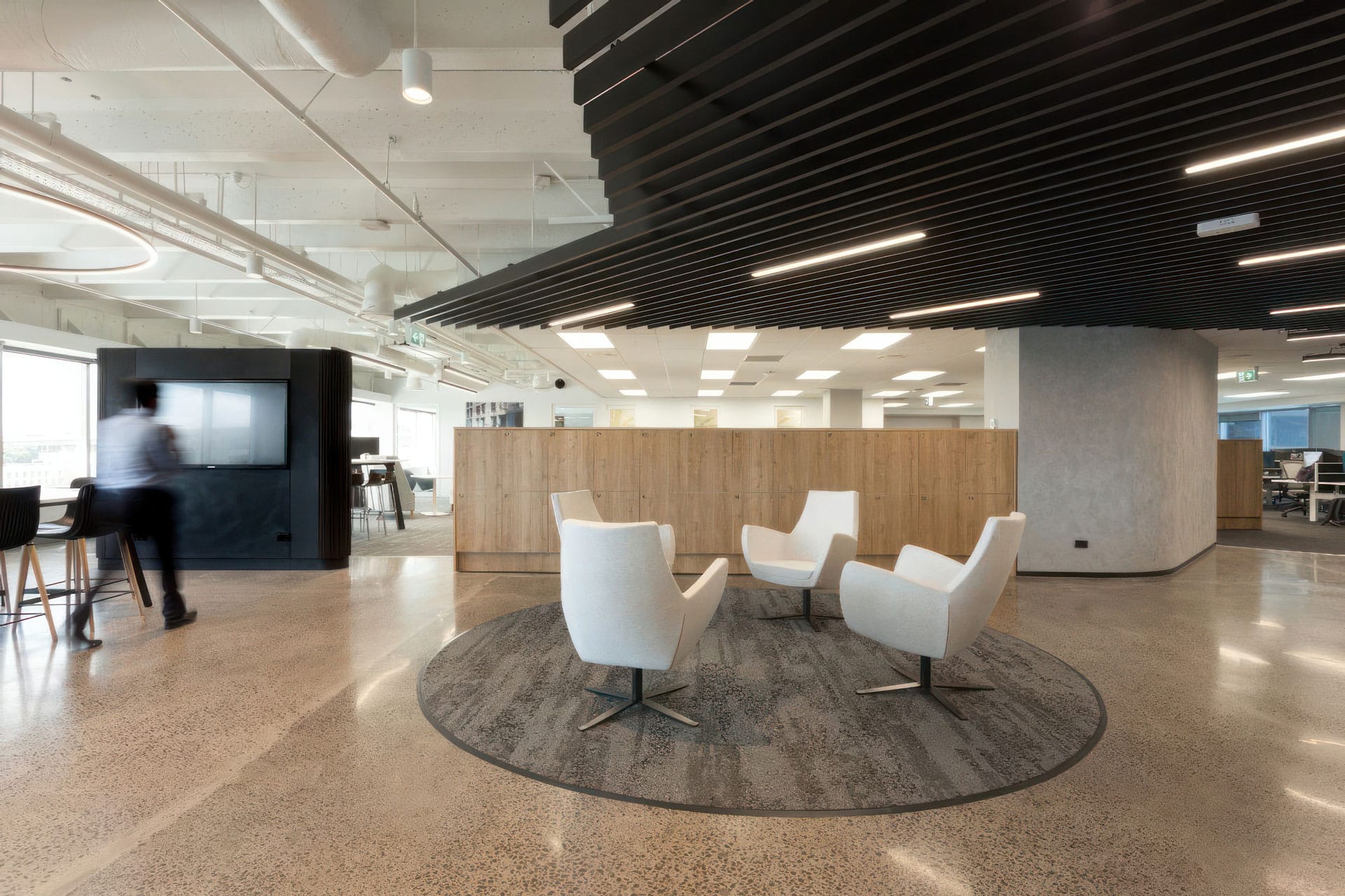 Modern office space with four white chairs arranged in a circle on a round rug, polished concrete floors, a wooden partition, and a person walking near a screen in the background. Ceiling features black slatted panels.