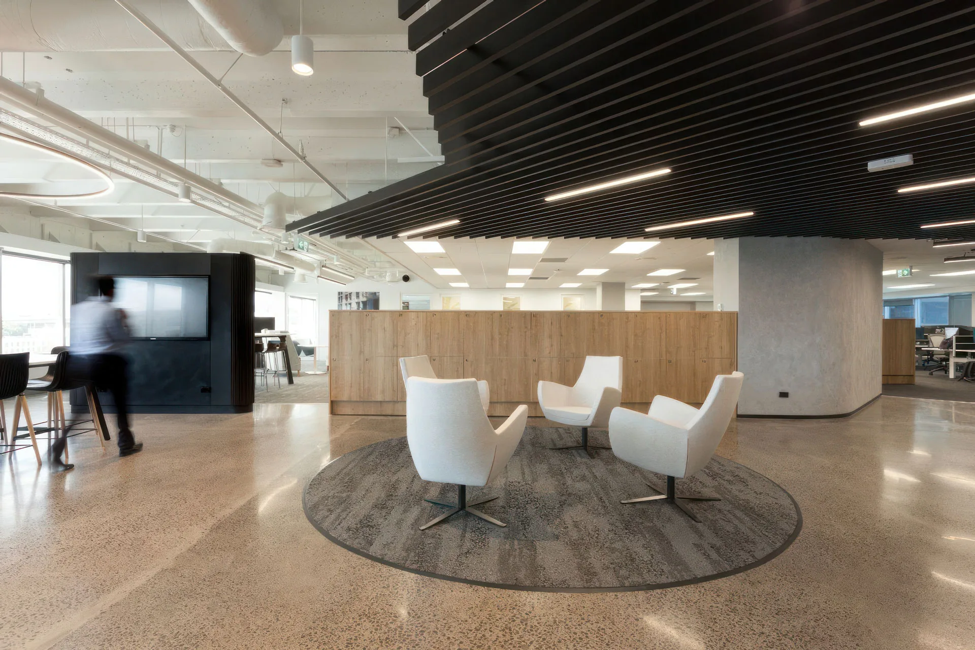 Modern office space with four white chairs arranged in a circle on a round rug, polished concrete floors, a wooden partition, and a person walking near a screen in the background. Ceiling features black slatted panels.
