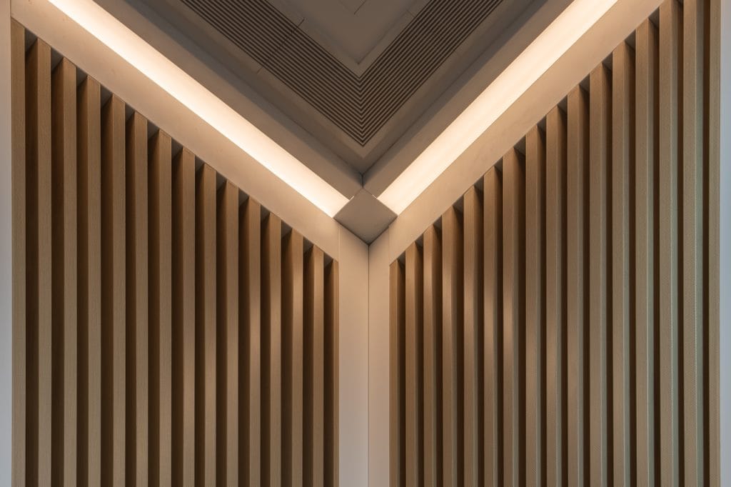 Symmetrical architectural ceiling design featuring vertical wooden slats converging at a corner, complemented by noise control with aluminium acoustic panels and soft, recessed lighting for clean, modern lines and a neutral color palette.