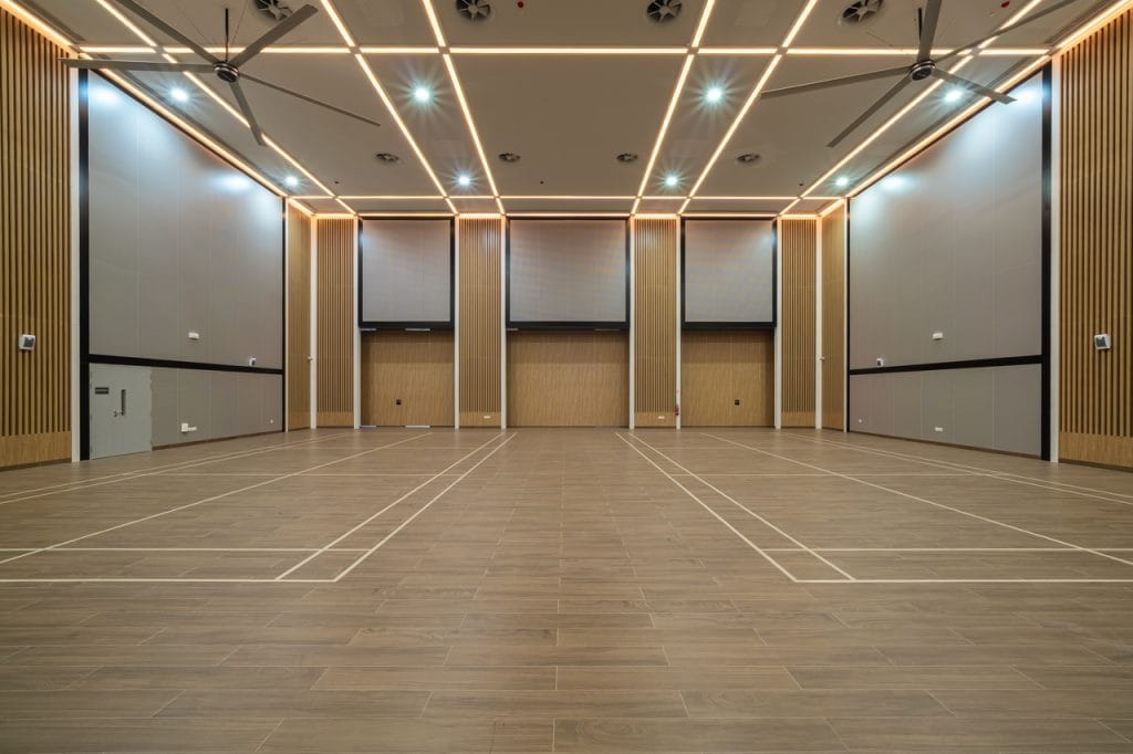 A large, empty indoor sports hall with wooden flooring, high ceilings, LED strip lights, and badminton court markings. The walls feature vertical wooden panels and Noise Control with Aluminium Acoustic Panels for a well-lit, acoustically balanced space.