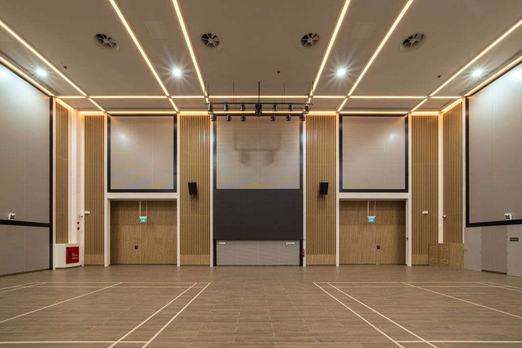 A spacious, modern indoor hall featuring high ceilings, wood paneling, Noise Control with Aluminium Acoustic Panels, LED strip lighting, a large projector screen at the front, and a court marked on the polished wooden floor.