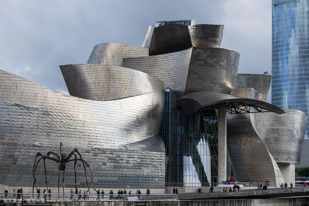 The Guggenheim Museum Bilbao, with its curving, aluminium exterior exemplifies Aluminium in Modern Architecture. A large spider sculpture stands before it, as people gather near the entrance and a modern glass building rises in the background.
