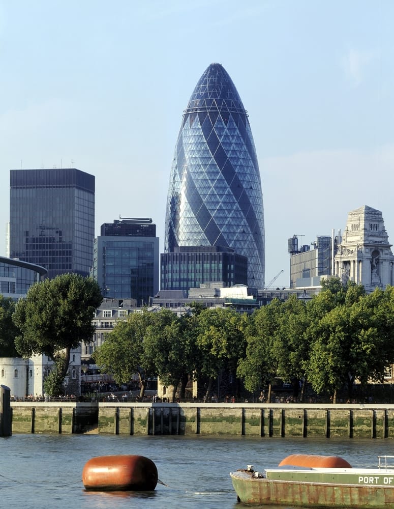 A view of London’s skyline featuring the glass-covered Gherkin building, showcasing aluminium in modern architecture, with trees in the foreground and the River Thames and a boat at the bottom of the image.