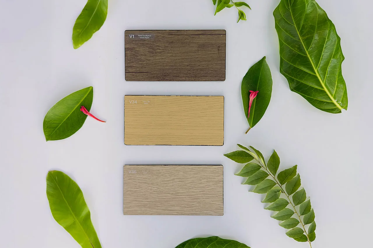 Three rectangular wood samples (brown, light tan, and beige) arranged vertically on a white surface, surrounded by various green leaves and two small pink flowers.
