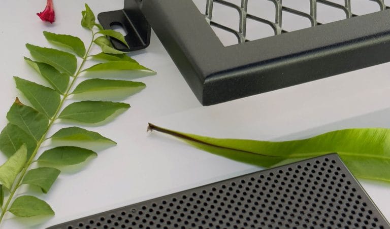 Close-up of green leaves and a small red flower next to two black, metal objects—one with a diamond mesh pattern and one with small circular holes—on a white surface.