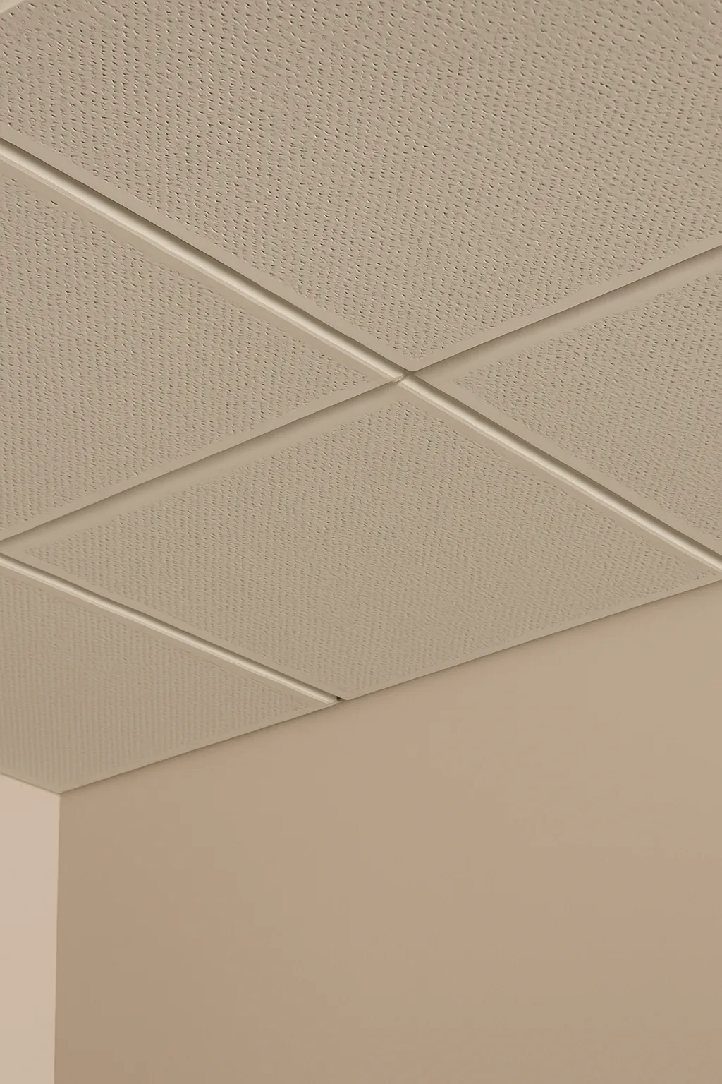A beige drop ceiling with textured acoustic tiles in a grid pattern above a cream-colored wall corner.