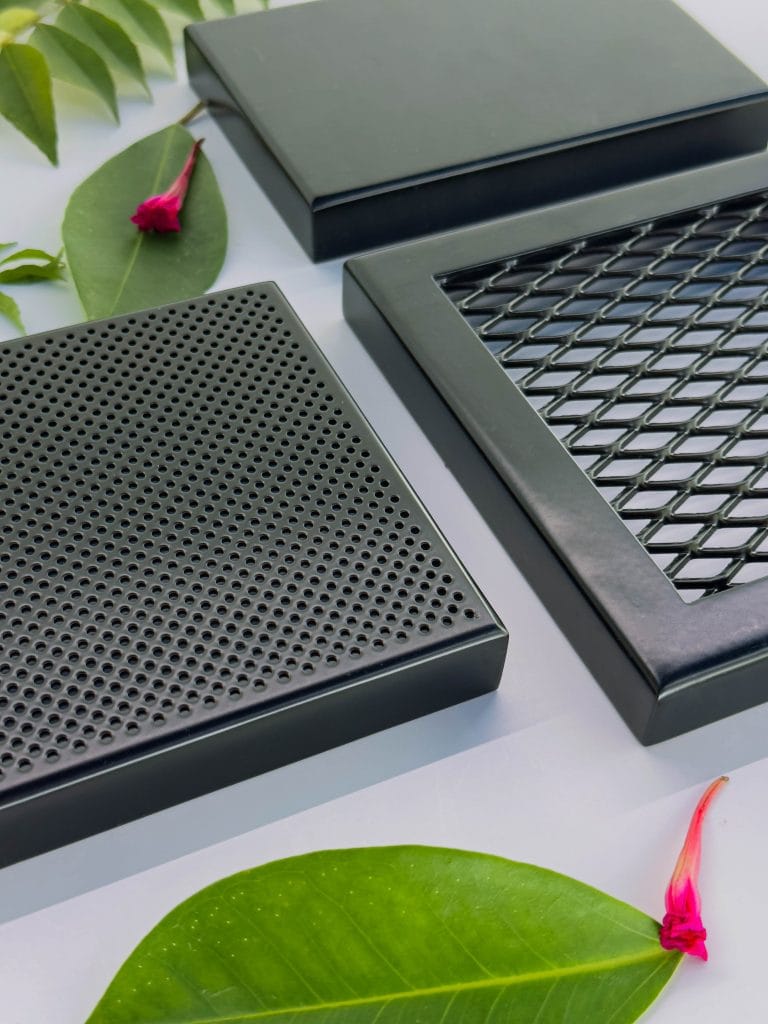 Three black metal vent or grille covers with different patterns—perforated holes, mesh, and diamond lattice—are arranged on a white surface, surrounded by green leaves and small pink flowers.