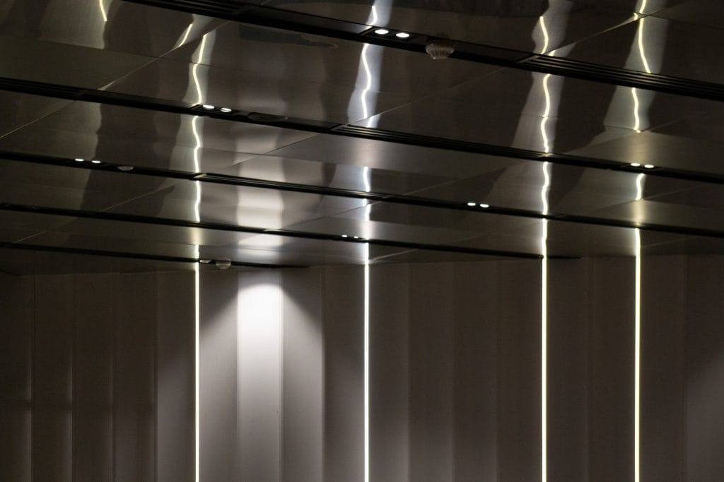 A modern ceiling with reflective metal panels and integrated lights, casting soft light and shadows on the plain, vertical wall panels below.