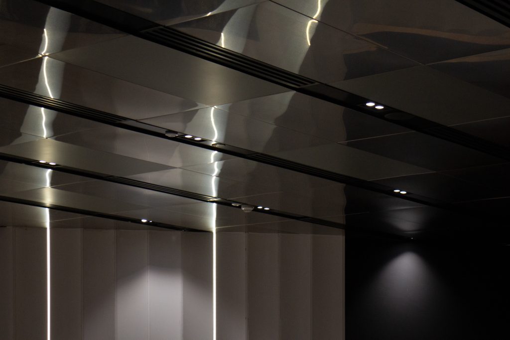 A modern ceiling with reflective metallic panels and built-in spotlights. Below, vertical white panels and a black wall are partially visible, with soft lighting creating a sleek, minimalist atmosphere.