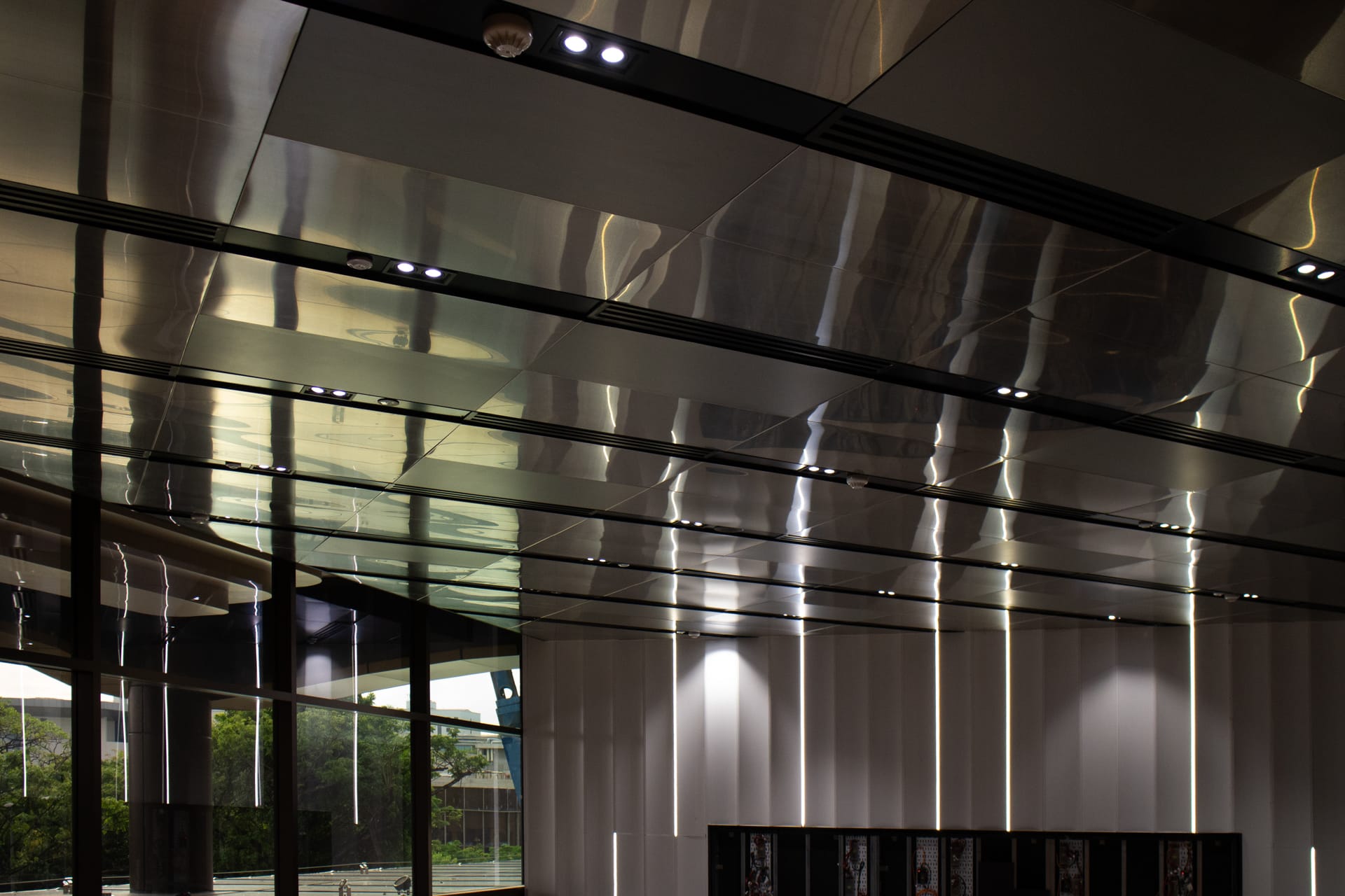 Modern interior ceiling with reflective metallic panels, recessed lights, and vertical light strips on the walls. Large windows reveal greenery and outdoor scenery, creating a sleek, contemporary atmosphere.