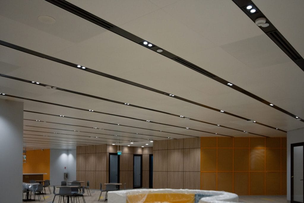 A modern office space with wood-paneled walls, yellow accent panels, recessed ceiling lights, scattered chairs, and tables. Some furniture is covered in protective plastic. The area looks clean and newly finished.