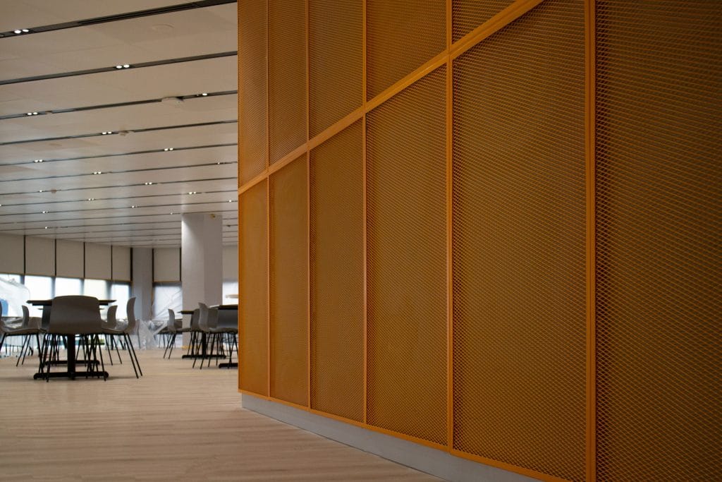 A modern interior space with light wooden floors, gray chairs around tables, and a prominent yellow perforated wall divider on the right. Ceiling lights create a clean, bright atmosphere.