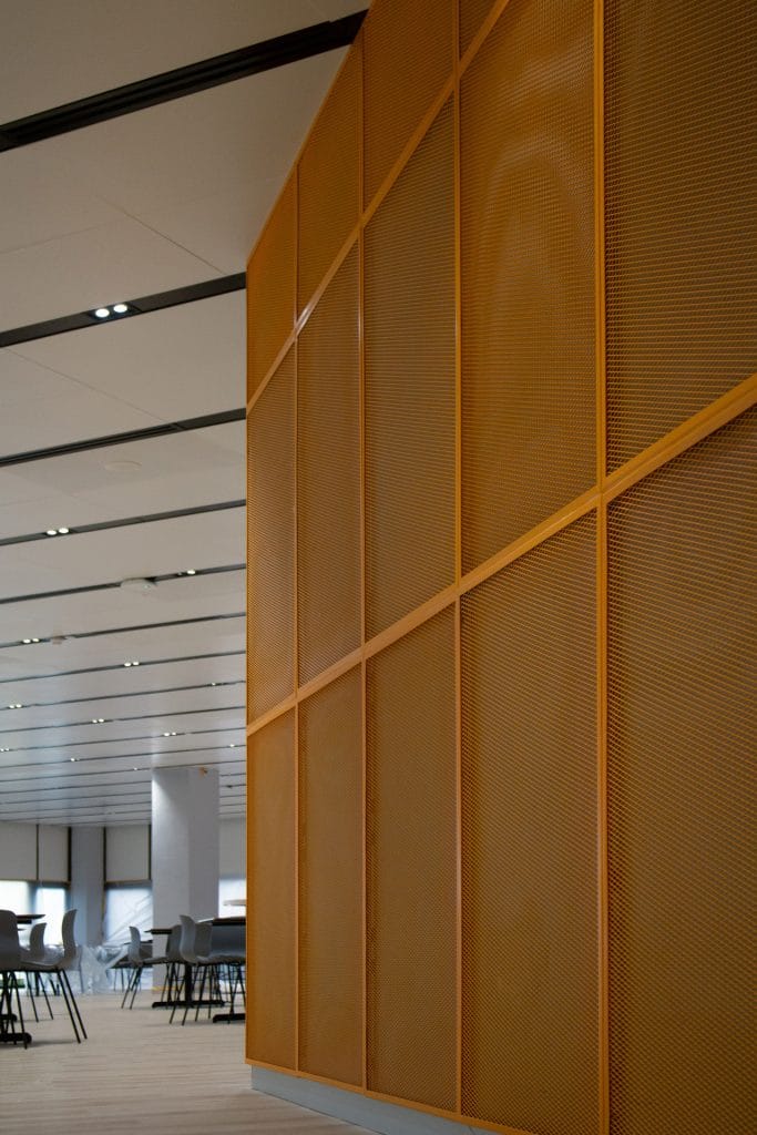 A modern interior with a tall orange perforated metal wall on the right, wooden flooring, and several tables and chairs arranged in the background under a white ceiling with recessed lights.