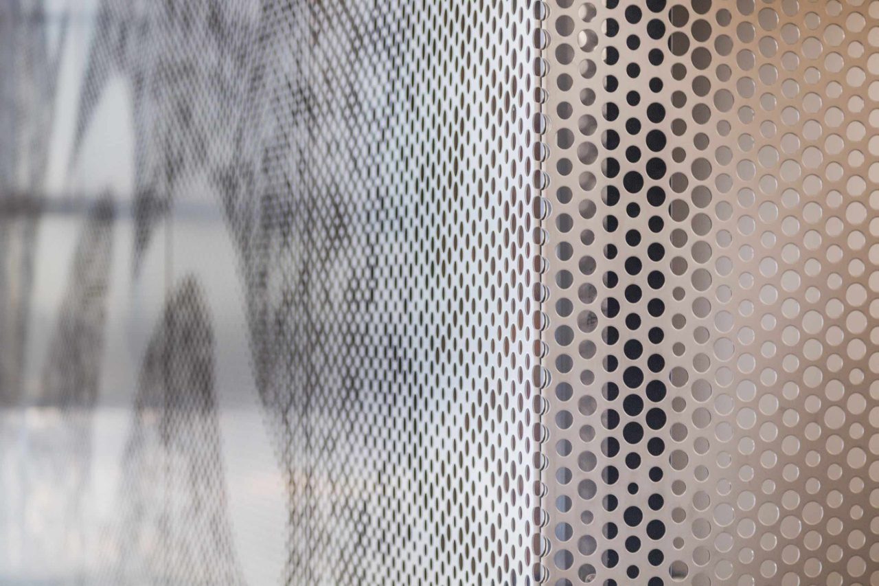 Close-up of two metal surfaces with perforated patterns. The left side shows a blurred, partially transparent sheet, while the right side displays a sharply focused beige sheet with evenly spaced round holes. Light shines through both.