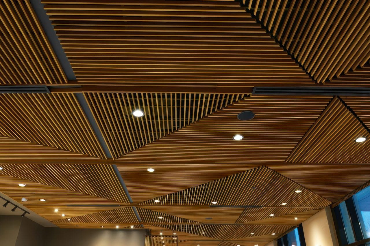 Ceiling with geometric wooden slats arranged in triangular and rectangular patterns, featuring recessed lights and linear light fixtures, creating a modern architectural design. The ceiling extends above a spacious interior.