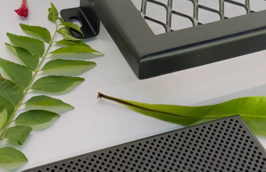 Close-up of green leaves and a small red flower next to two black, metal objects—one with a diamond mesh pattern and one with small circular holes—on a white surface.