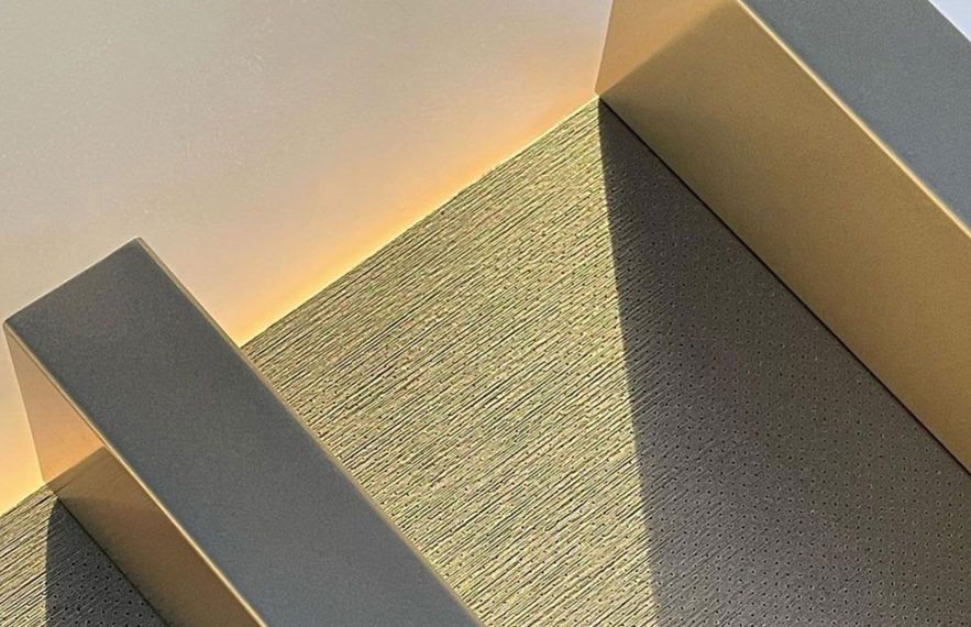 Sunlight casts geometric shadows on a modern staircase with textured steps and smooth railings, creating a minimalist and abstract pattern with soft yellow lighting on the wall.