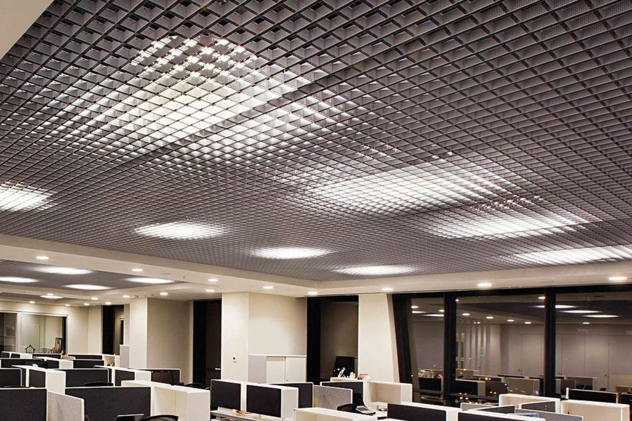 Modern office interior with an open layout, featuring rows of cubicles and workstations. The ceiling has a grid design with built-in fluorescent lights, creating a geometric pattern. Large windows line one wall.
